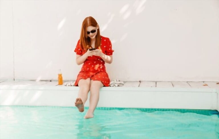 Beautiful young redhead woman wearing red dress using mobile phone while resting by the swimming pool Beautiful young redhead woman wearing red dress using mobile phone while resting by the swimming pool