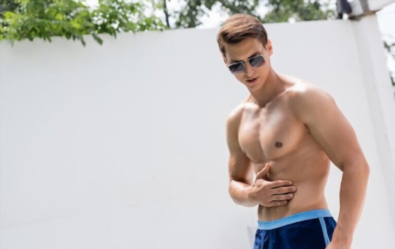 man in sunglasses and swimming trunks applying sunscreen on muscular torso man in sunglasses and swimming trunks applying sunscreen on muscular torso