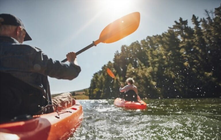 Outdoor photo of senior man rowing boat in lake with woman in background on a summer day. Men and women in two different kayaks in the lake on a sunny day. Outdoor photo of senior man rowing boat in lake with woman in background on a summer day. Men and women in two different kayaks in the lake on a sunny day.