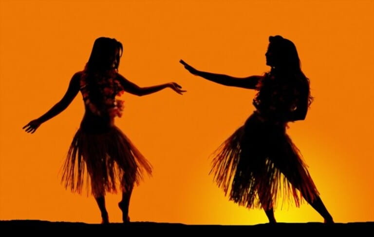 A silhouette of two women dancing in their grass skirts, in the outdoors.