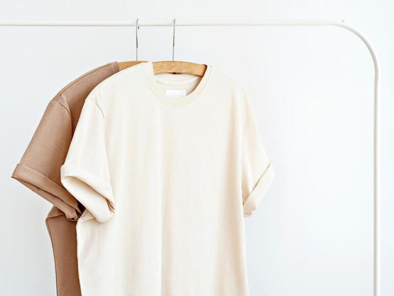 Two t-shirts in neutral milky colours hanging on white rack against white wall. Blank template, mockup for textile design. Summer minimalist outfit idea. Capsule wardrobe. Two t-shirts in neutral milky colours hanging on white rack against white wall. Blank template, mockup for textile design. Summer minimalist outfit idea. Capsule wardrobe.