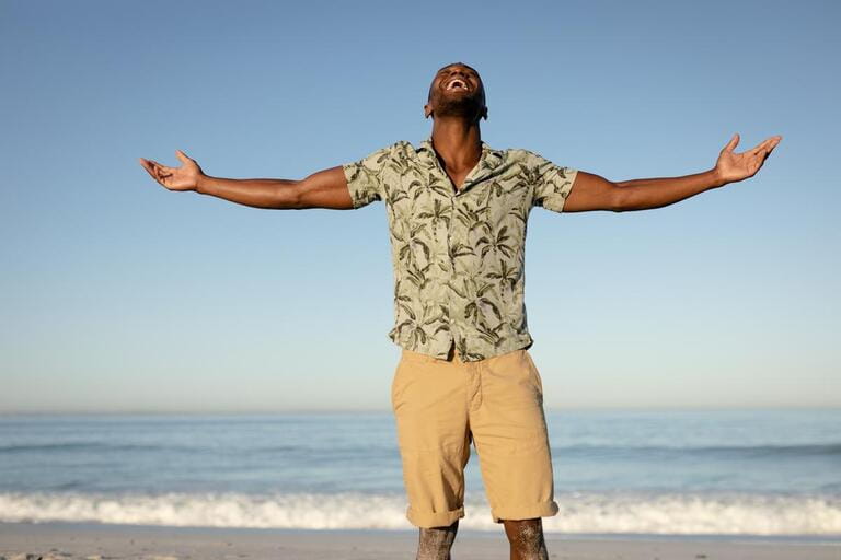 A happy, attractive African American man enjoying free time on beach on a sunny day, wearing a Hawaiian shirt, sun shining on his face with his arms outstretched. Relaxing summer vacation. A happy, attractive African American man enjoying free time on beach on a sunny day, wearing a Hawaiian shirt, sun shining on his face with his arms outstretched. Relaxing summer vacation.
