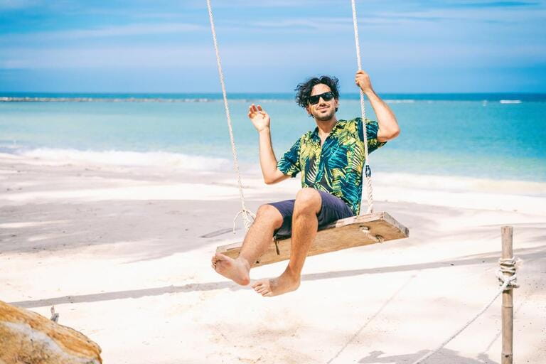 Stylish happy sexy young brunette man with a beard in a Hawaiian shirt is swinging on a swing on the tropical sea