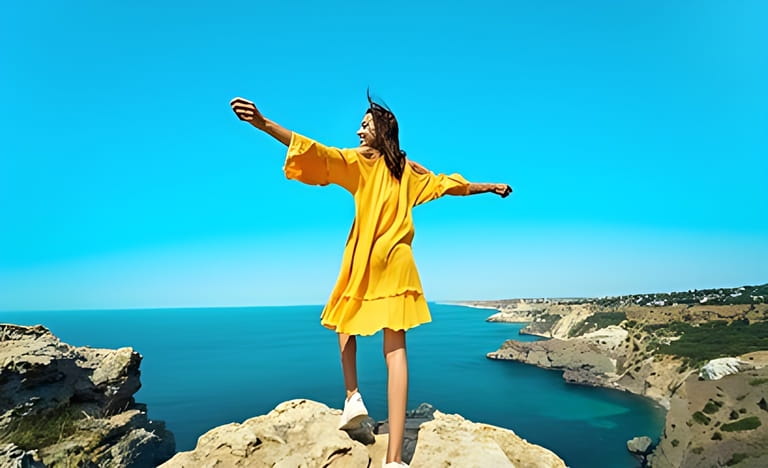 Rear view of tanned traveler woman standing on top rock beach with open arms in front of amazing seascape, bright yellow dress and hair blowing in the wind. Rear view of tanned traveler woman standing on top rock beach with open arms in front of amazing seascape, bright yellow dress and hair blowing in the wind.